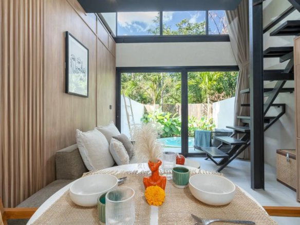 Modern dining area with a woven table runner, white bowls, and teal glassware; wooden wall panels and a metal staircase visible through large windows with greenery outside.