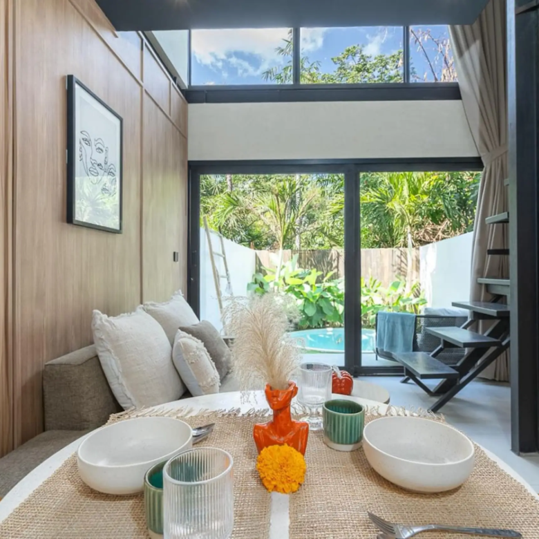 Dining table set with white bowls, green cups, and orange flower centerpieces in a bright, modern living space with wooden wall panels and a glass door to a garden.