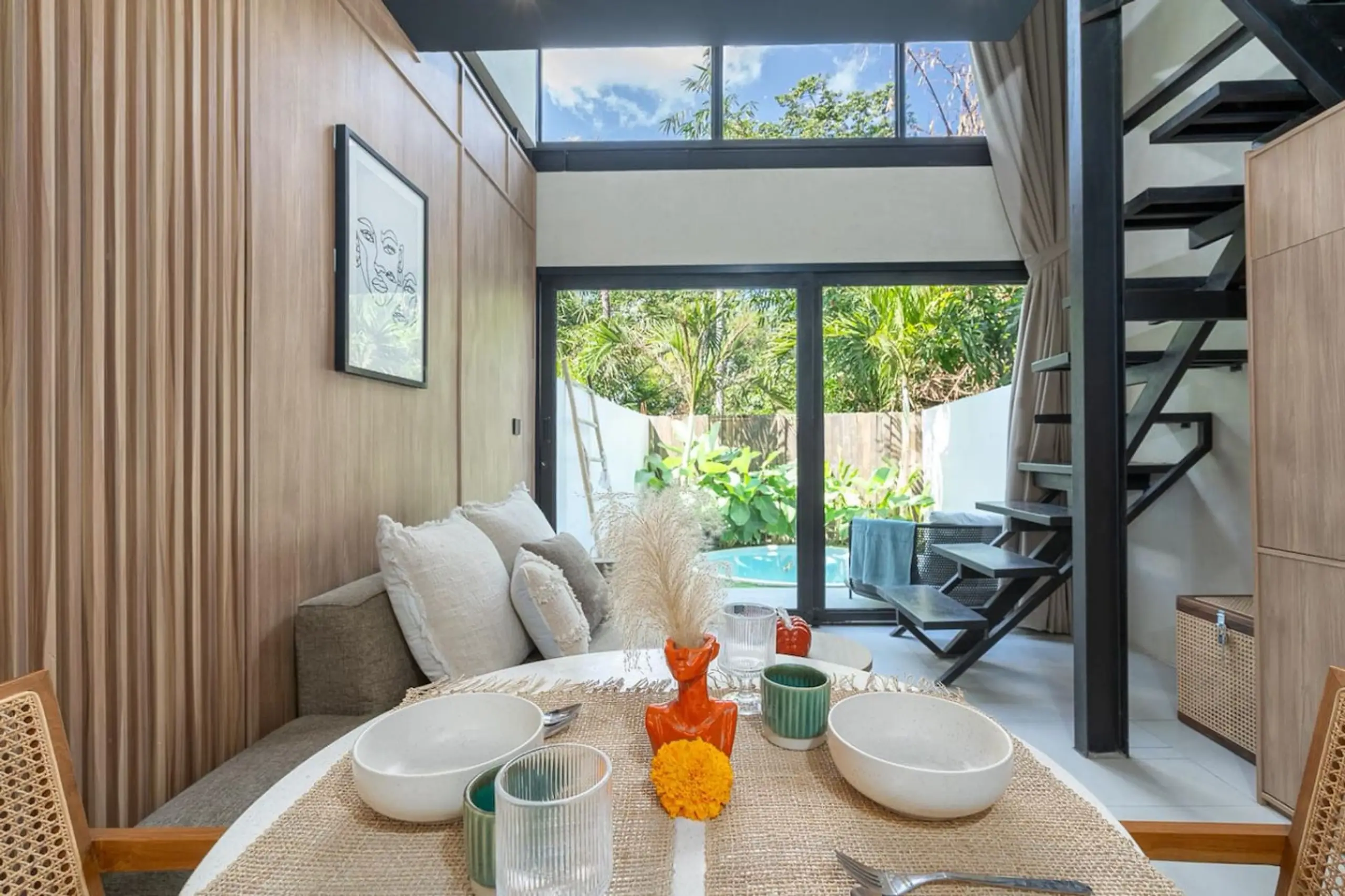 Dining table set with white bowls, green cups, and orange flower centerpieces in a bright, modern living space with wooden wall panels and a glass door to a garden.