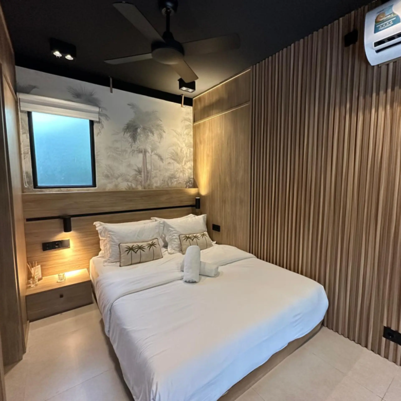 Bedroom with two white beds, Palm tree-patterned pillows, wooden wall panels, and warm bedside lighting.