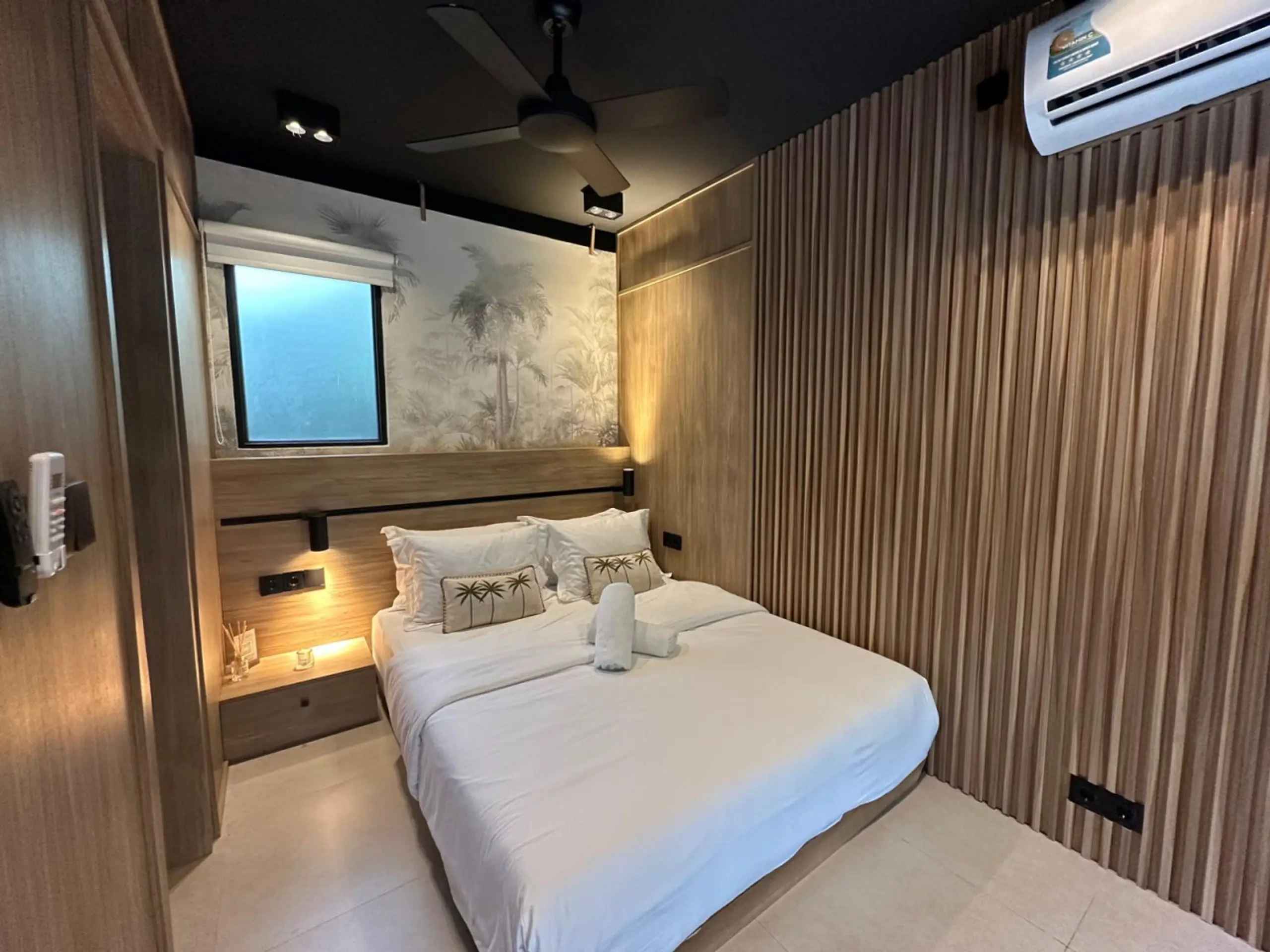 Bedroom with two white beds, Palm tree-patterned pillows, wooden wall panels, and warm bedside lighting.