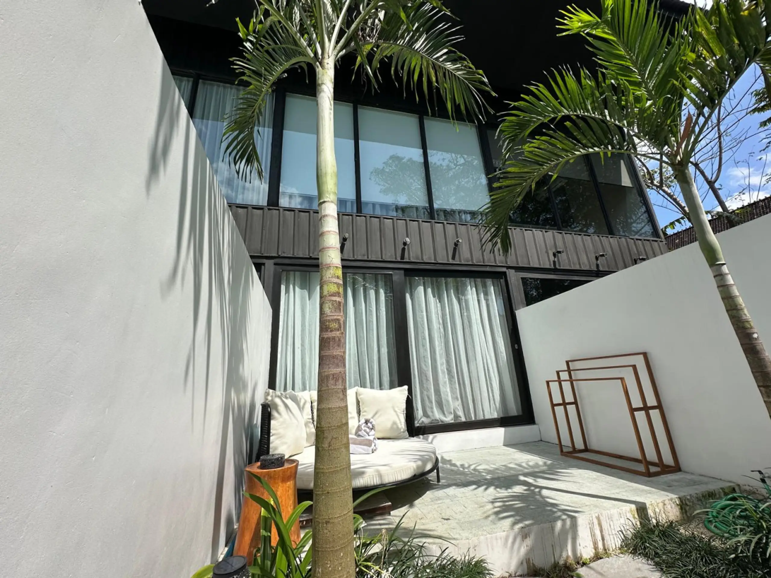 Modern courtyard with white walls, palm trees, and a glass-fronted contemporary building, plus cushioned seating by the wall.
