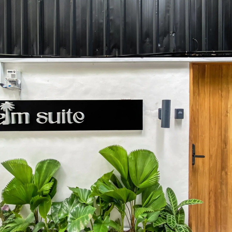 Indoor wall scene with a black 'pam suite' sign, lush tropical plants in front, and a wooden door on the right.