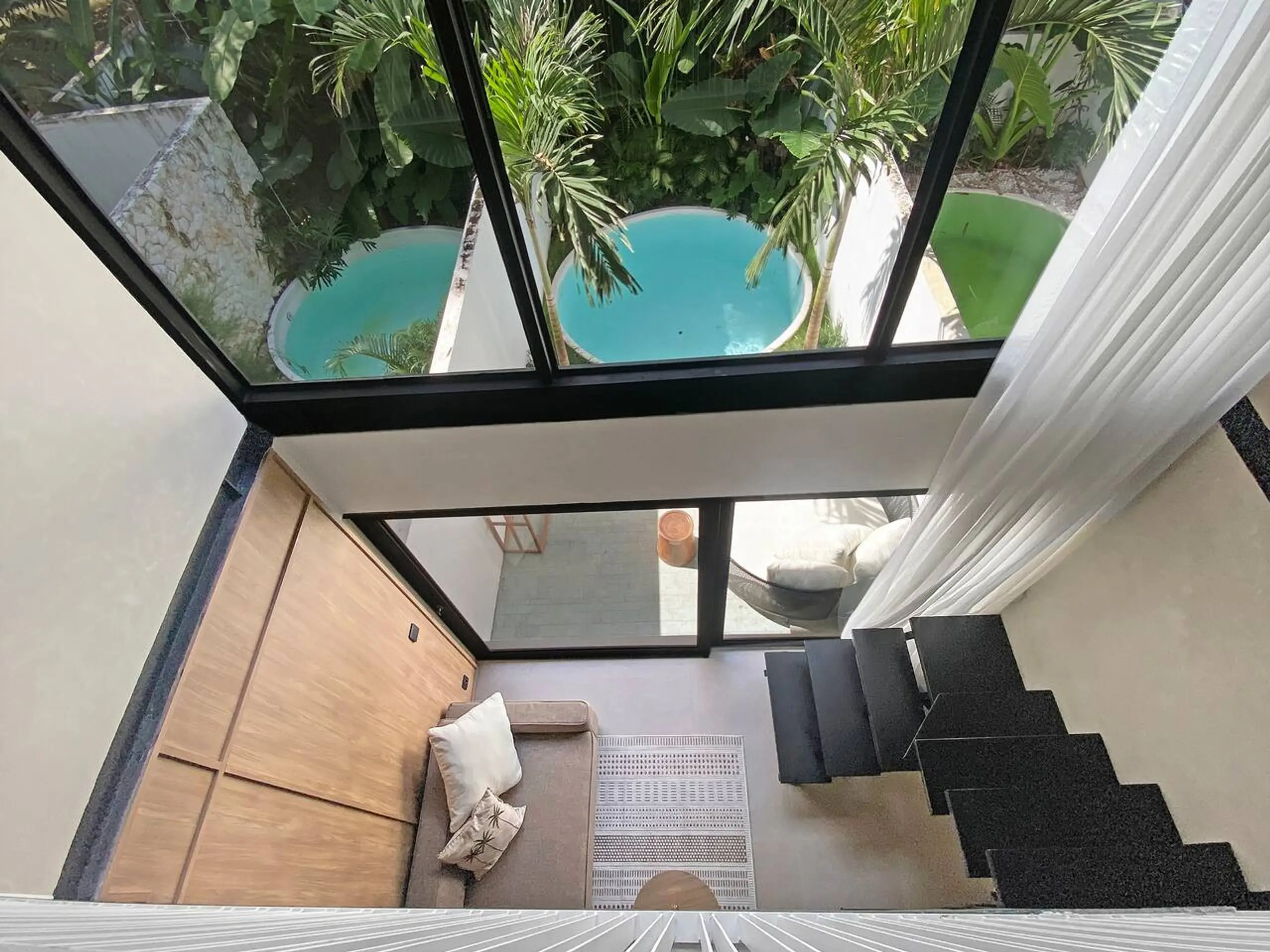Modern stairwell and seating area seen through large glass walls with tropical plants outside.
