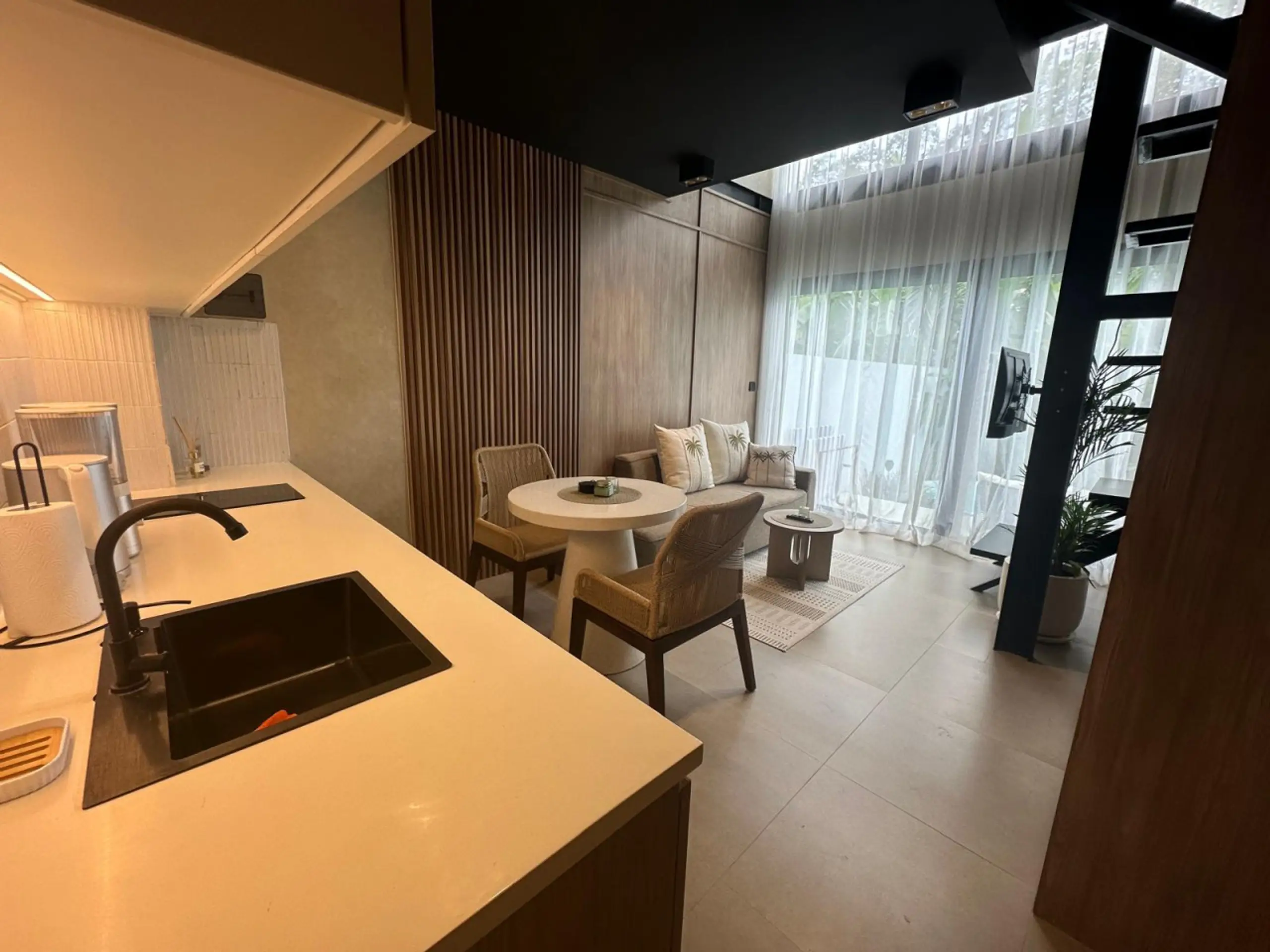 Contemporary open-plan kitchen and living area with an island, sink, and pillowed seating by large windows with sheer curtains.
