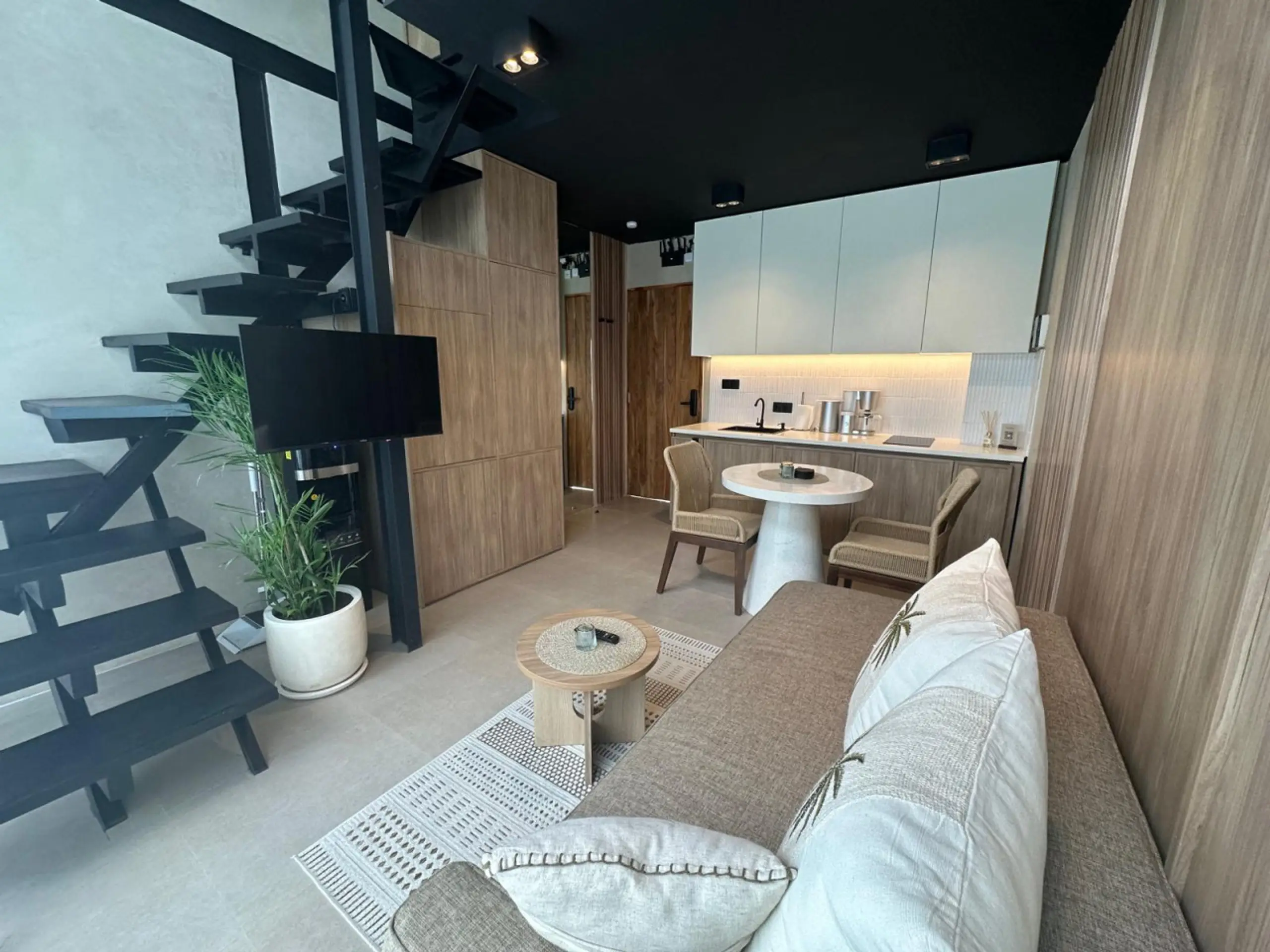 Modern living area with a black metal staircase, wood cabinets, white countertops, and a beige sofa with throws; green plant in a white pot nearby.