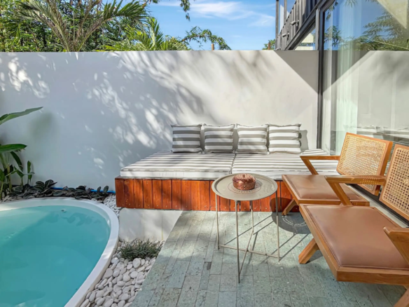 Outdoor poolside seating area with striped cushions on a wooden bench, a round metal table, and wicker chairs under a sunny sky.