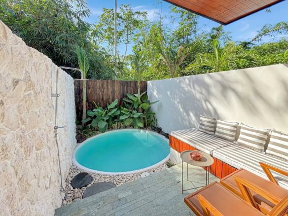 Small round turquoise pool in a tropical courtyard with a stone wall, wooden fence, and striped lounge chairs nearby.