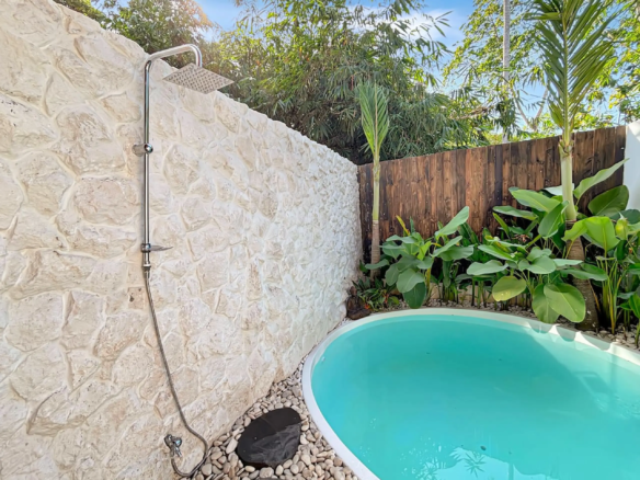 Outdoor shower mounted on a textured stone wall beside a curved turquoise pool, with tropical plants and a wooden fence in the background.