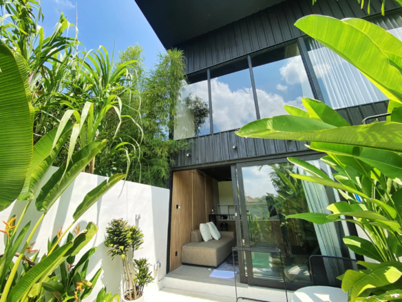 Modern house with dark metal siding and tall glass windows surrounded by dense tropical plants in a bright outdoor courtyard.