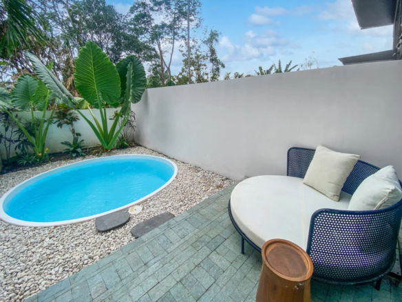 Backyard pool area with a small oval blue pool, pebble ground, wooden stools, and cushioned wicker chairs against a tall white wall with tropical plants nearby.