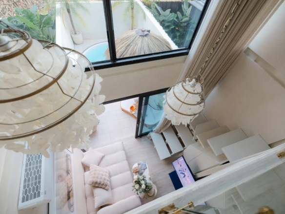 Living room seen from above: white sectional sofa, glass doors, and hanging chandeliers with tropical plants outside.