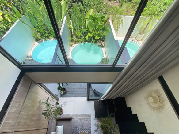 Indoor stairwell with a glass ceiling revealing a courtyard pool and tropical plants beneath a sunny sky at center stage.