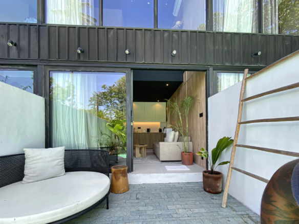 Contemporary outdoor patio with curved white seating, black wicker chair, glass doors, and potted plants.