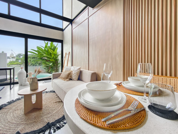 Bright contemporary dining area with round white tables set with bowls, plates, and glassware, a beige sofa, and a wooden slat wall beside large glass windows with greenery outside.