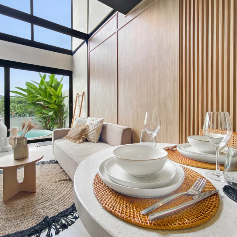 Bright contemporary dining area with round white tables set with bowls, plates, and glassware, a beige sofa, and a wooden slat wall beside large glass windows with greenery outside.