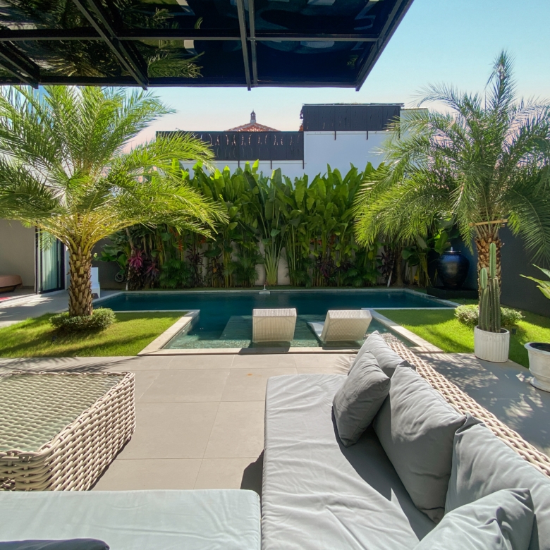 Modern backyard pool area with beige tiled deck, woven lounge chairs with gray cushions, and lush palm trees surrounding a rectangular pool.