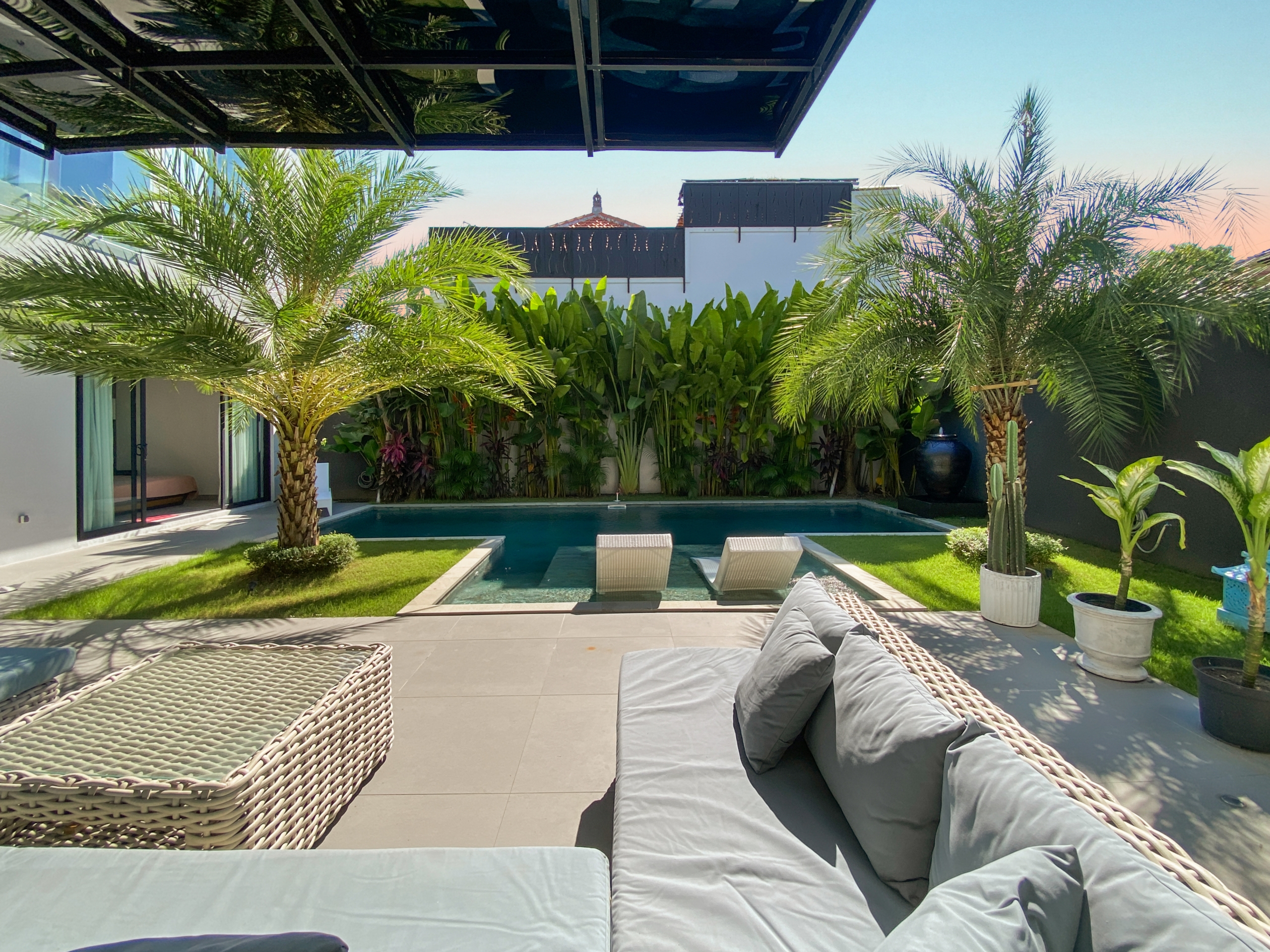 Modern backyard pool area with beige tiled deck, woven lounge chairs with gray cushions, and lush palm trees surrounding a rectangular pool.