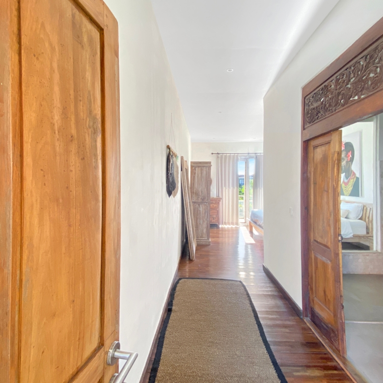 Narrow home hallway with tall wooden doors on both sides, white walls, and a beige runner rug leading to a sunlit room at the end.