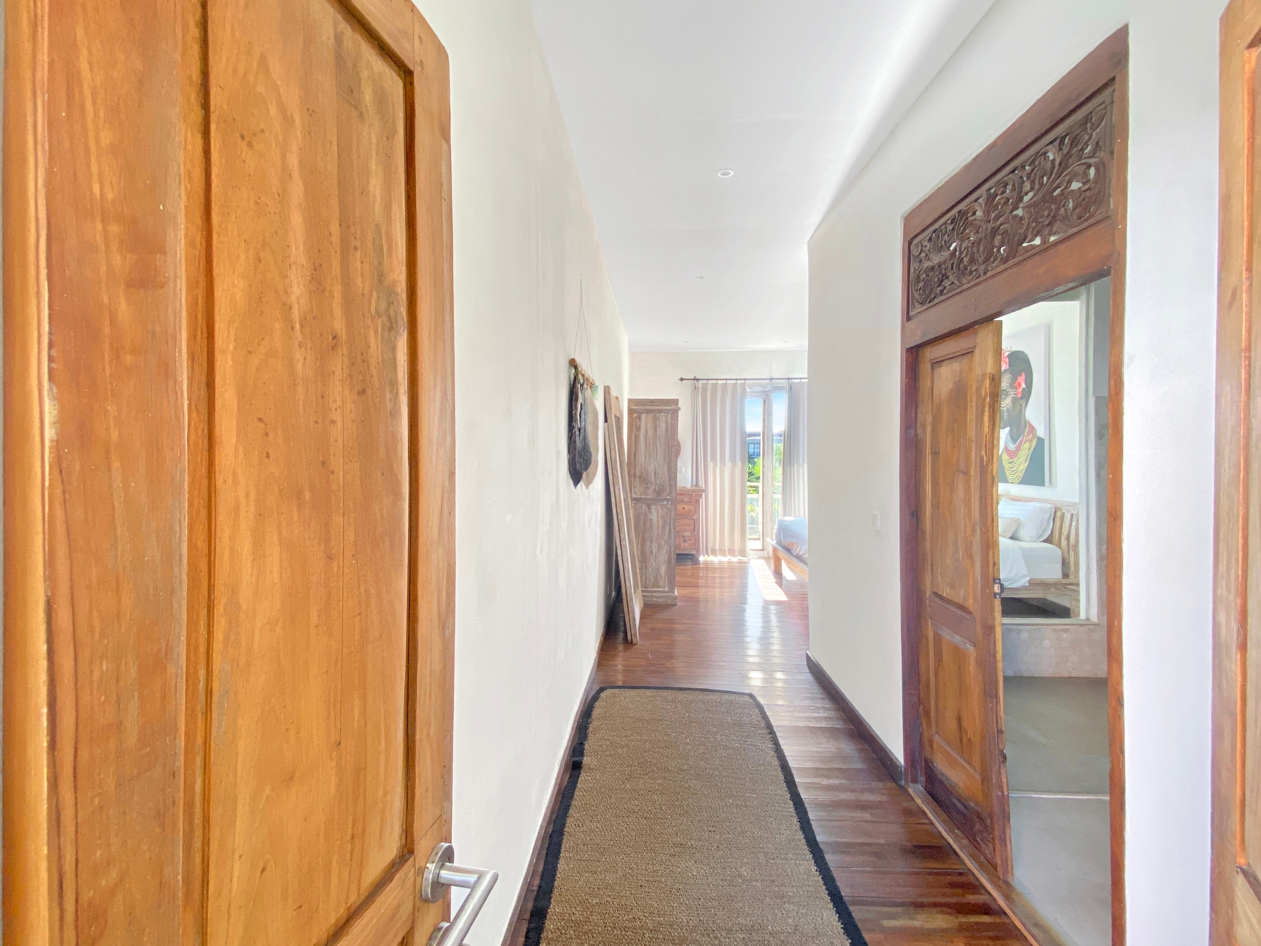 Narrow home hallway with tall wooden doors on both sides, white walls, and a beige runner rug leading to a sunlit room at the end.
