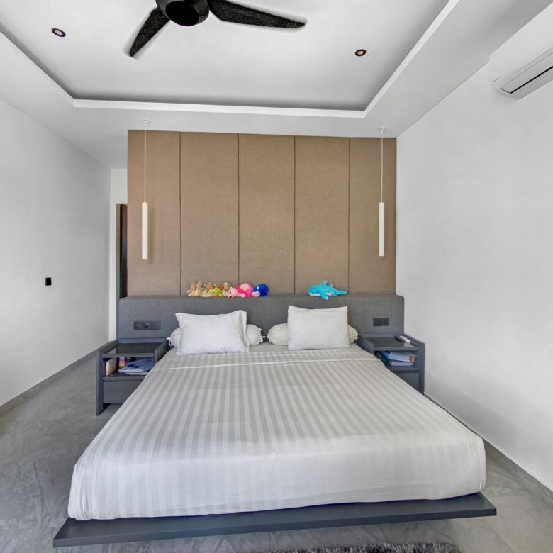 Modern minimalist bedroom with a low platform bed, grey-striped duvet, and a tall wooden headboard panel; pendant lights overhead and toys on the headboard shelf.