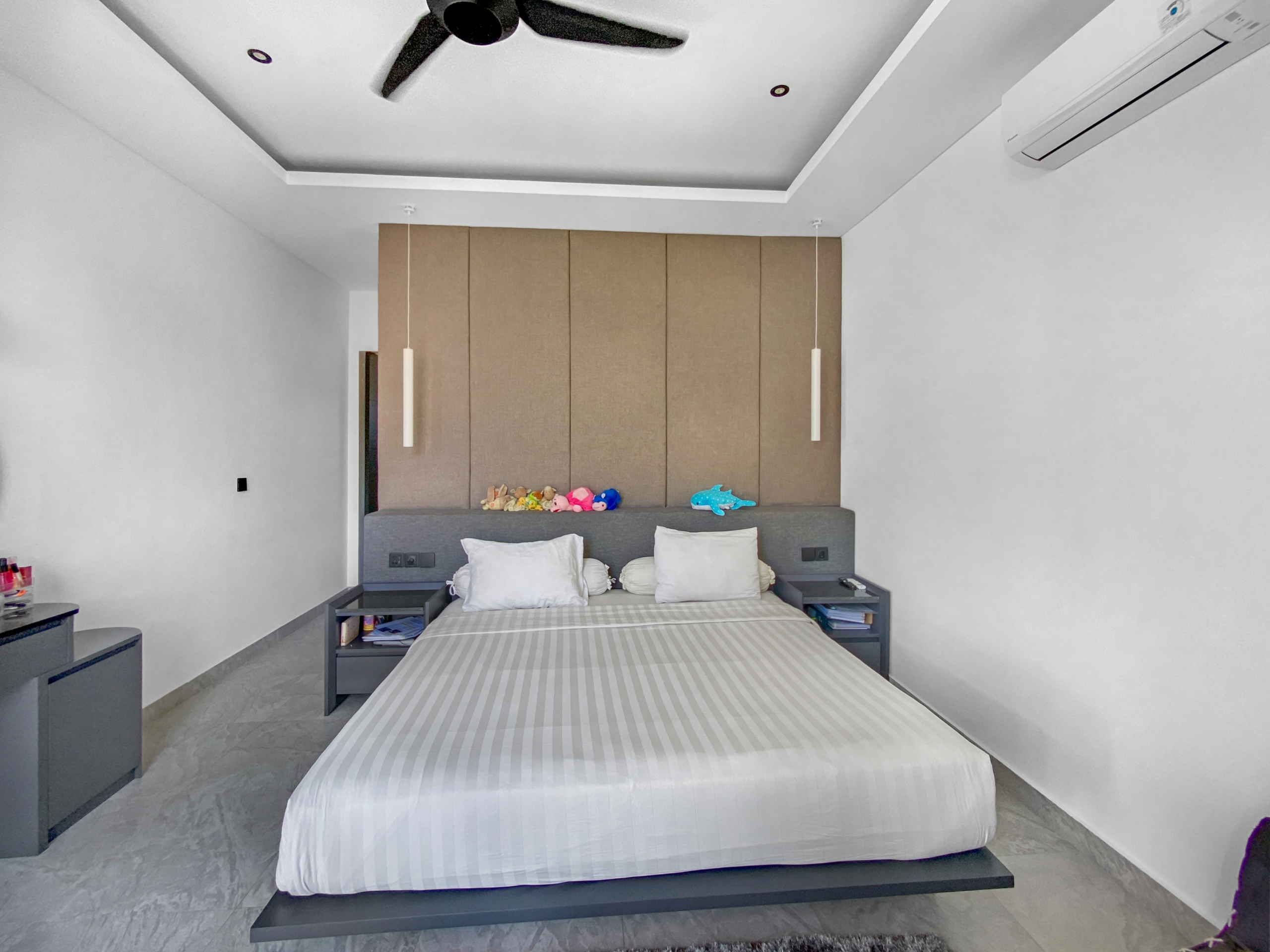 Modern minimalist bedroom with a low platform bed, grey-striped duvet, and a tall wooden headboard panel; pendant lights overhead and toys on the headboard shelf.