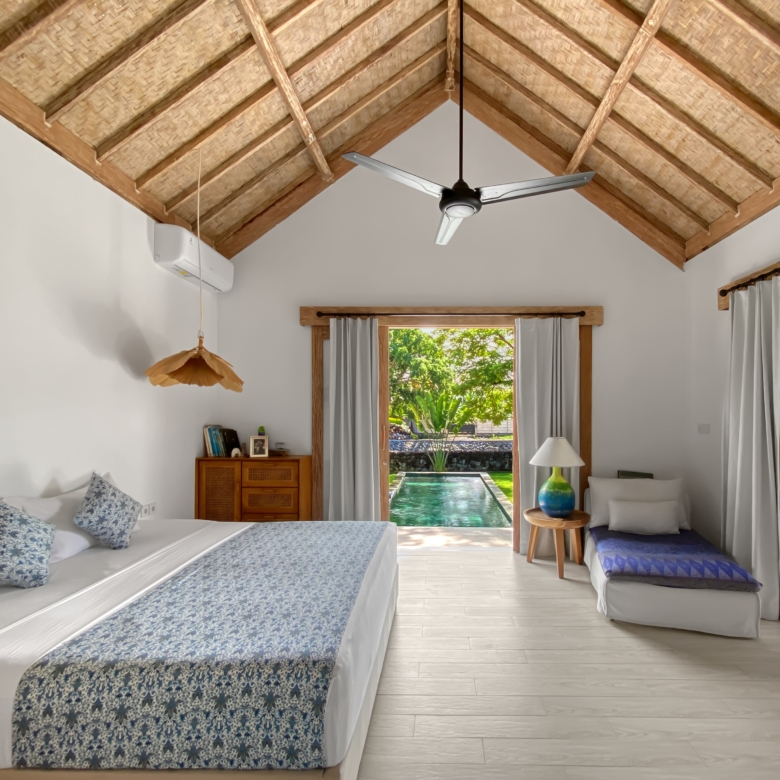 Room with a vaulted thatched ceiling and a ceiling fan, two white beds with blue patterned runners, and a doorway opening to a pool and garden.
