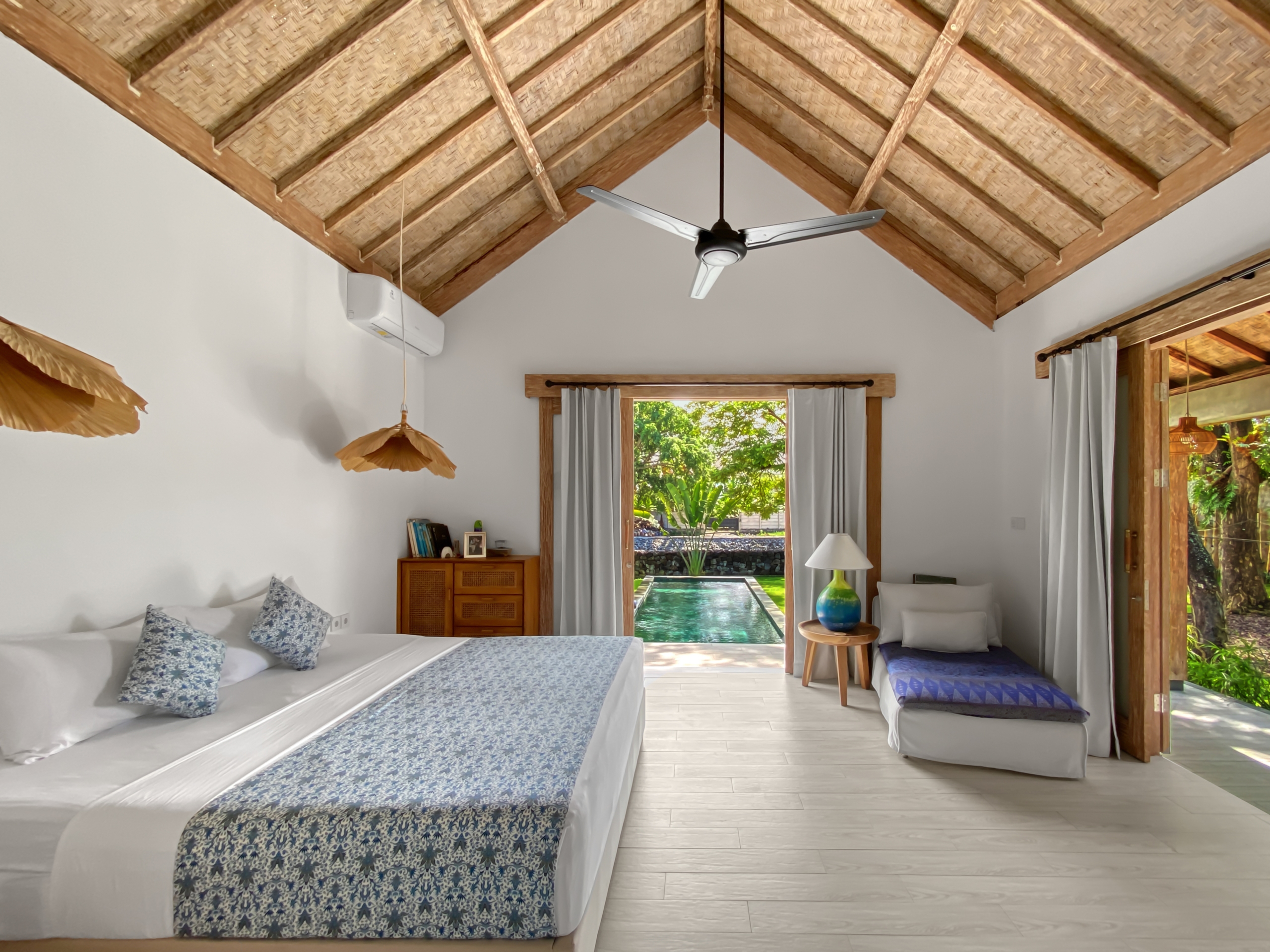08aa33d790b03d2ccbac9bf14ad3096b3c497715 Room with a vaulted thatched ceiling and a ceiling fan, two white beds with blue patterned runners, and a doorway opening to a pool and garden.