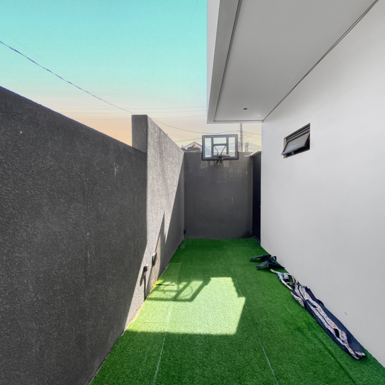 Narrow outdoor alley with green artificial grass, gray left wall and white right wall, and a distant basketball hoop under a low overhang.