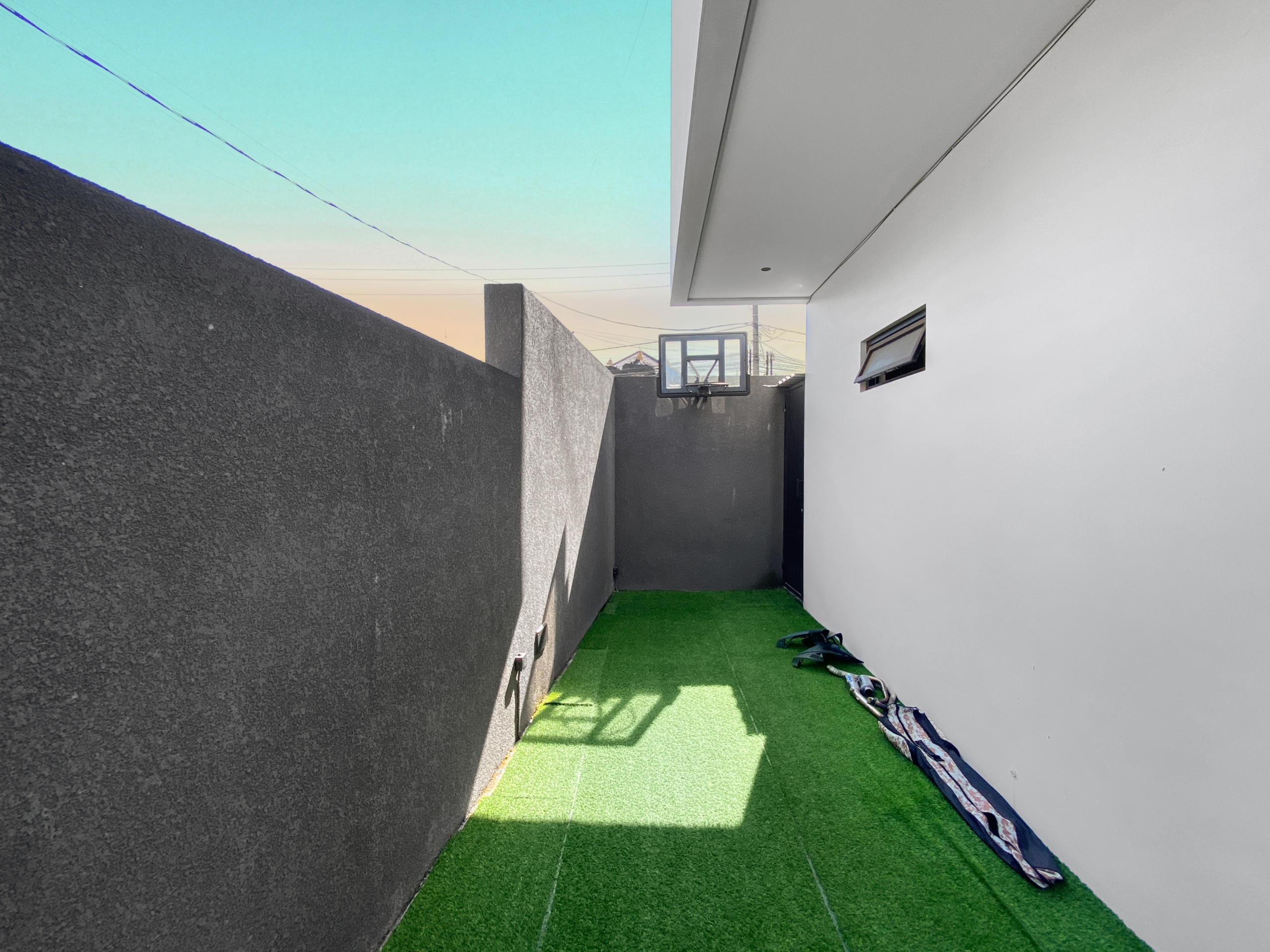 Narrow outdoor alley with green artificial grass, gray left wall and white right wall, and a distant basketball hoop under a low overhang.