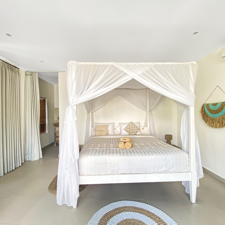 Bright bedroom with a white four-poster bed and sheer canopy, beige linens, and circular braided rugs on a tiled floor.