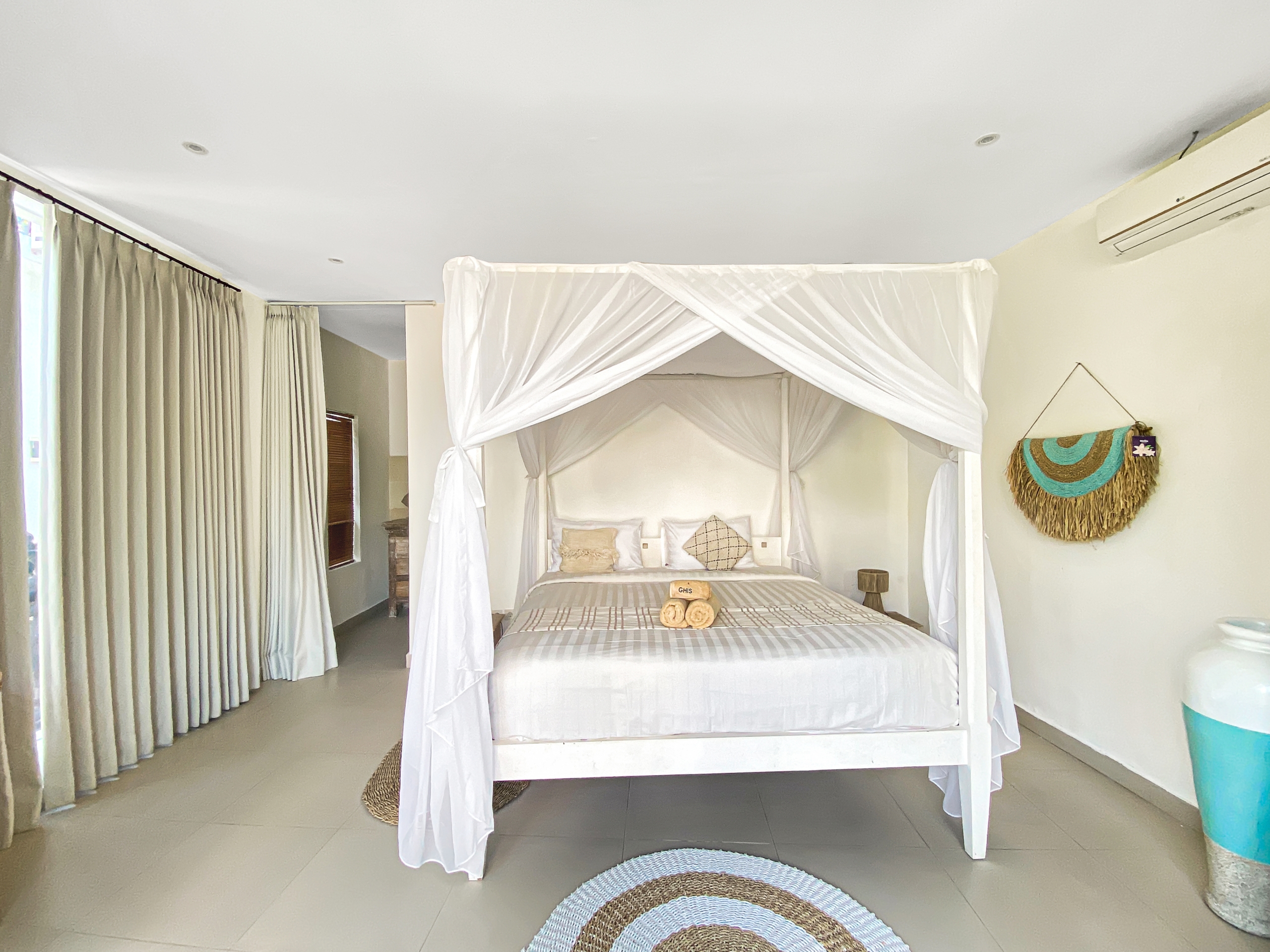Bright bedroom with a white four-poster bed and sheer canopy, beige linens, and circular braided rugs on a tiled floor.