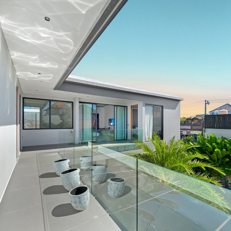 Modern balcony with a glass railing, gray tile floor, and white exterior walls; several white ceramic planters arranged along the edge, with a blue sky and neighborhood houses in the background.