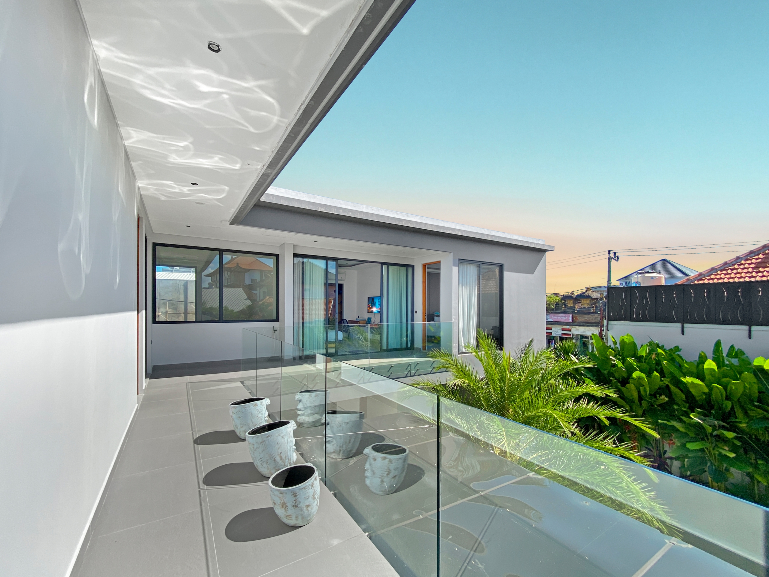 Modern balcony with a glass railing, gray tile floor, and white exterior walls; several white ceramic planters arranged along the edge, with a blue sky and neighborhood houses in the background.
