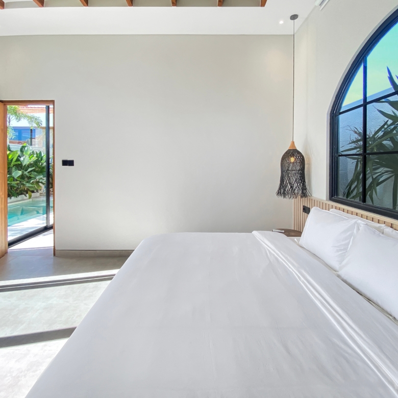 Bright hotel-style bedroom with white bedding, a wooden headboard, and a large arched window revealing outdoor plants.
