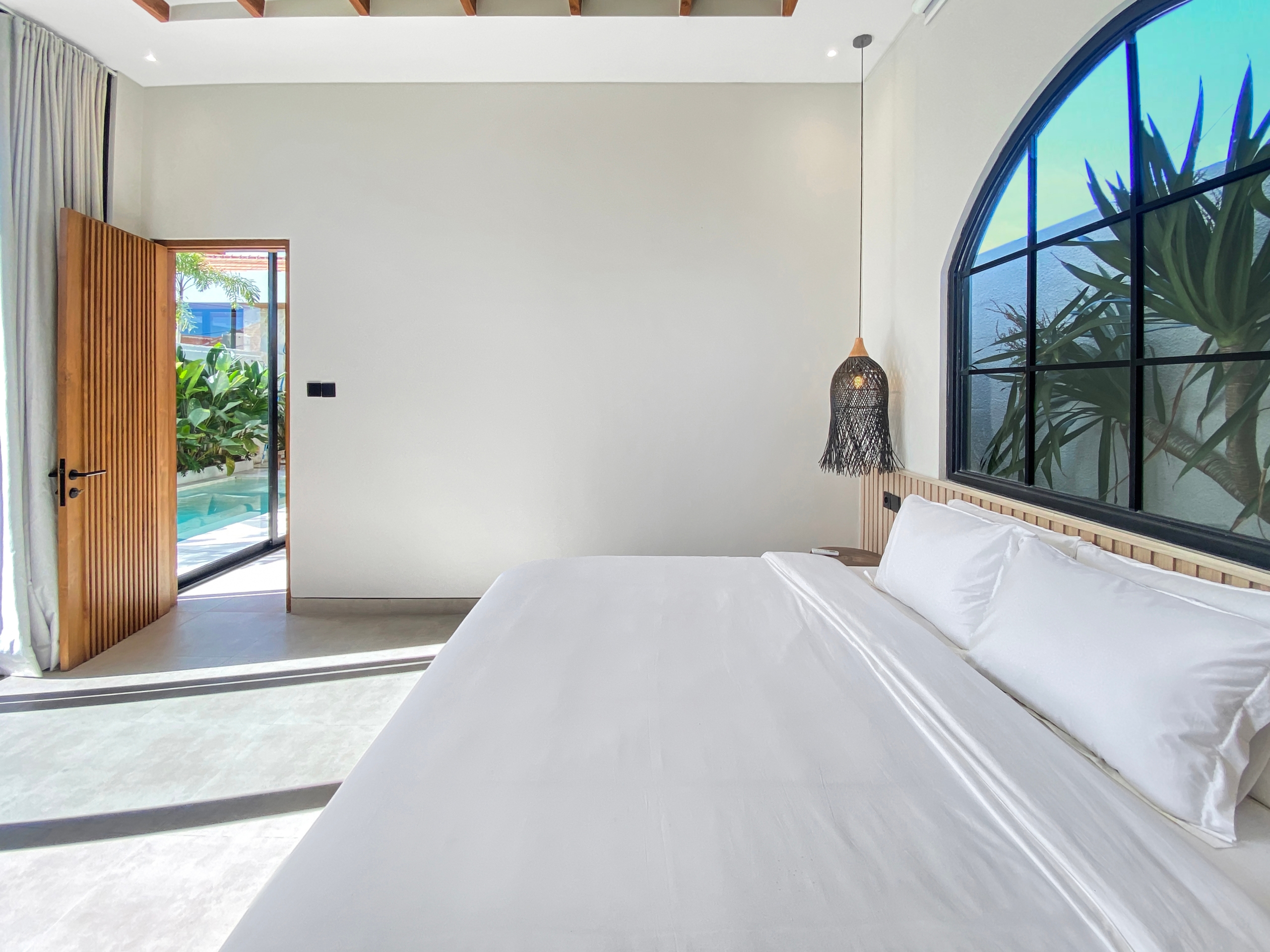 Bright hotel-style bedroom with white bedding, a wooden headboard, and a large arched window revealing outdoor plants.