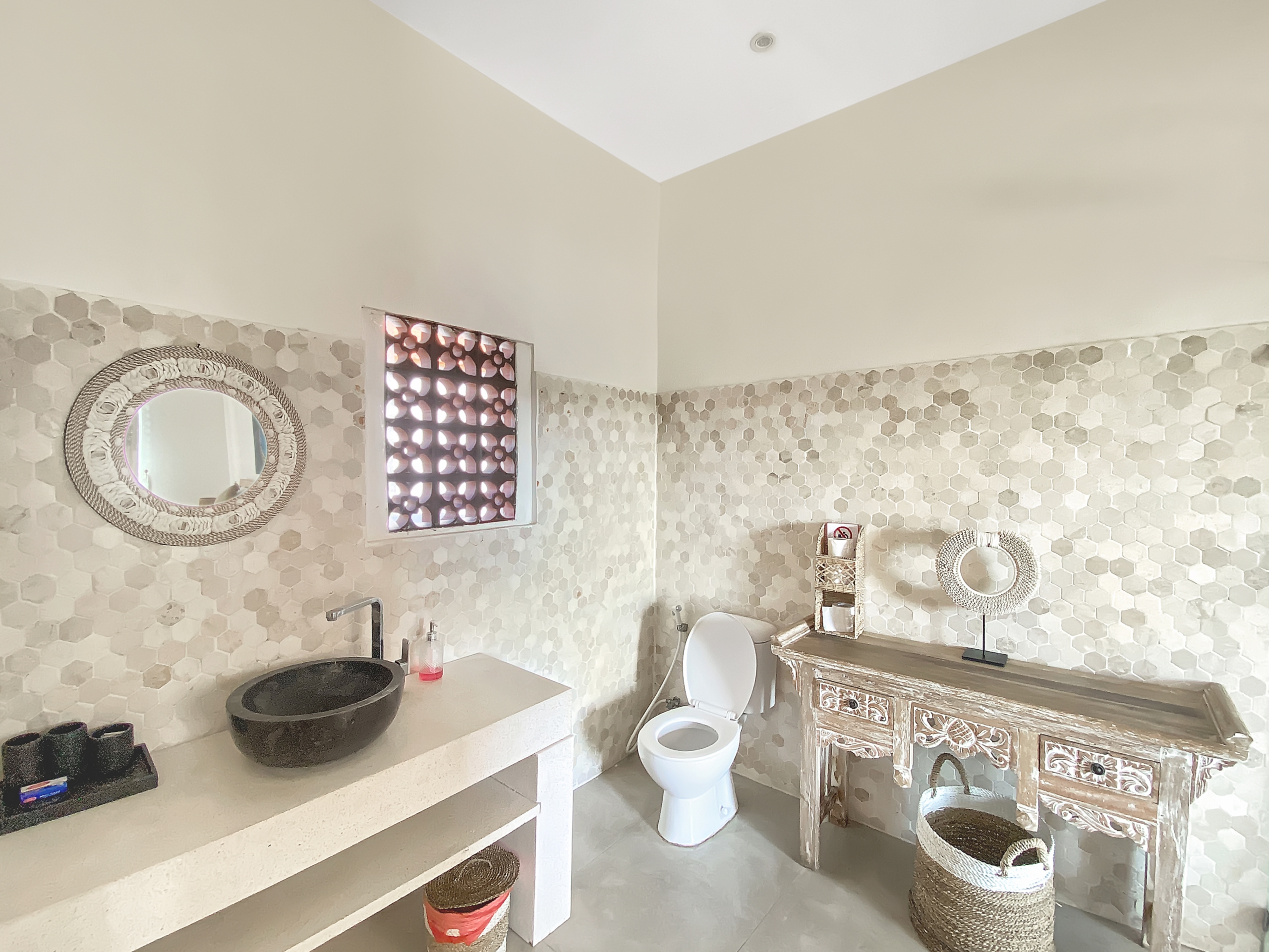 Bathroom with beige hex tile walls, round wicker-framed mirror, open vanity counter, and white toilet.