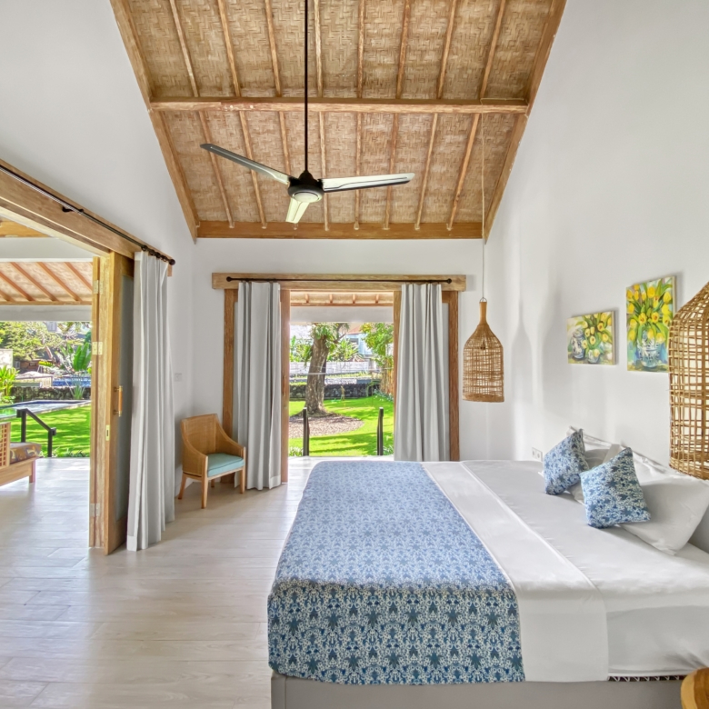 Bright tropical bedroom with two white beds, blue patterned runners, wicker pendant lamps, and a thatched ceiling; garden visible through open doors.