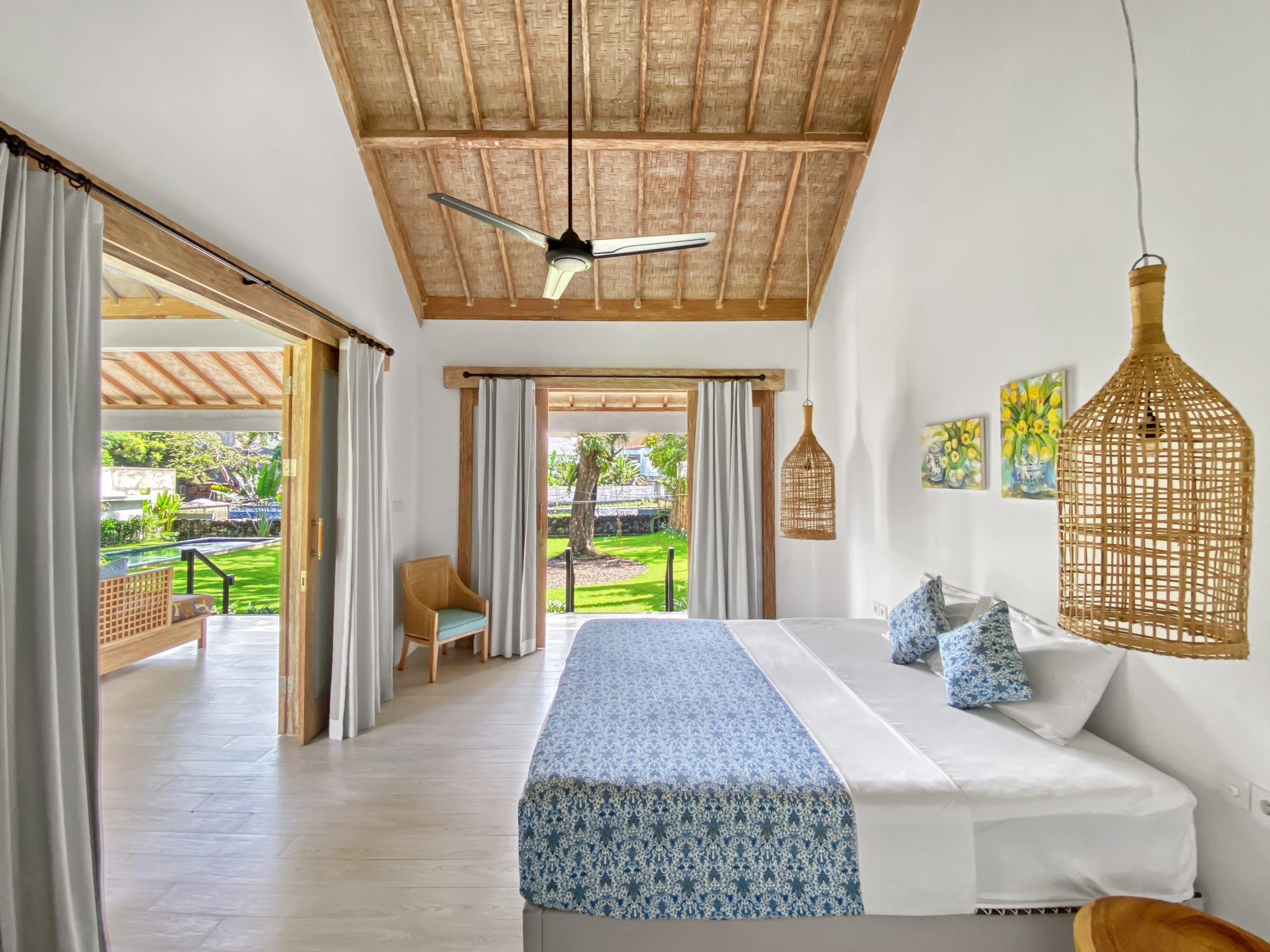 17731014c7442aa91f3ac885e80ed509f6be8c85 Bright tropical bedroom with two white beds, blue patterned runners, wicker pendant lamps, and a thatched ceiling; garden visible through open doors.