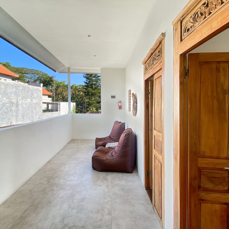 Covered balcony with white walls, tiled floor, brown cushioned seating, and carved wooden door frames; sunny outdoor scene with blue sky and greenery beyond.