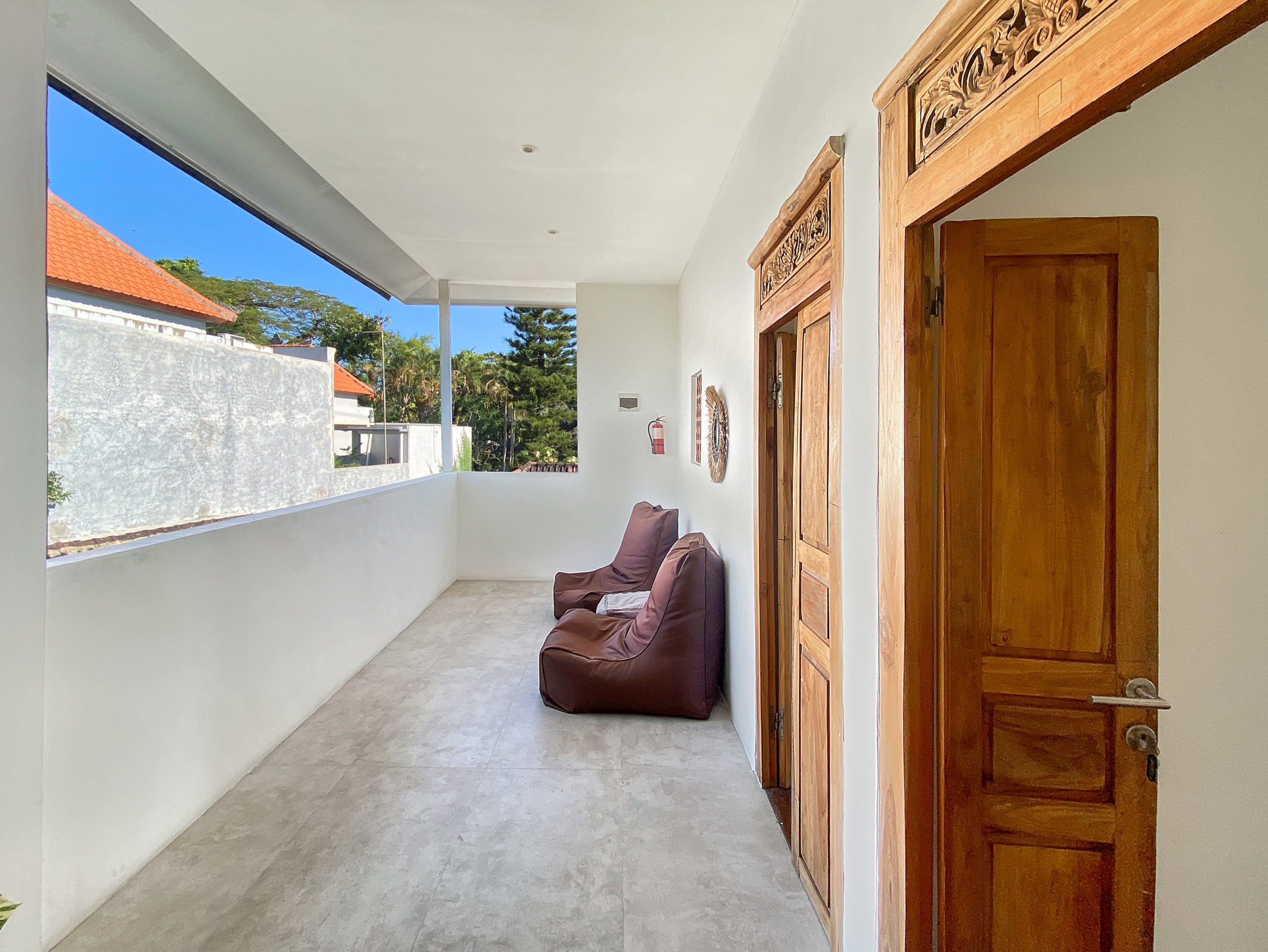 Covered balcony with white walls, tiled floor, brown cushioned seating, and carved wooden door frames; sunny outdoor scene with blue sky and greenery beyond.
