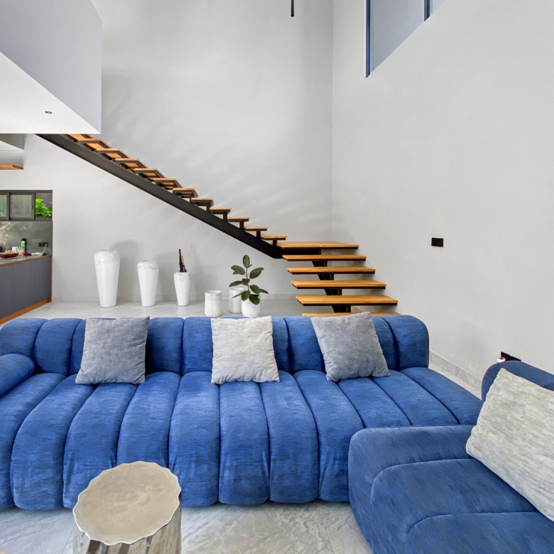 Bright living room with a large blue sectional sofa, gray cushions, and a floating wooden staircase against white walls.