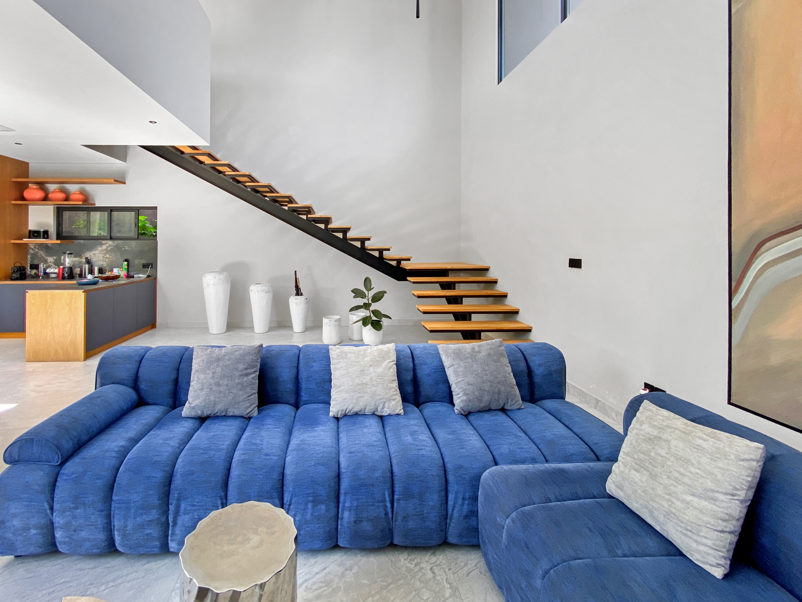Bright living room with a large blue sectional sofa, gray cushions, and a floating wooden staircase against white walls.
