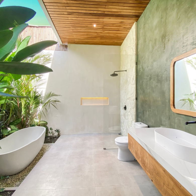Modern bathroom with a white oval freestanding tub on a wooden base, surrounded by lush tropical plants and a light gray tiled floor.