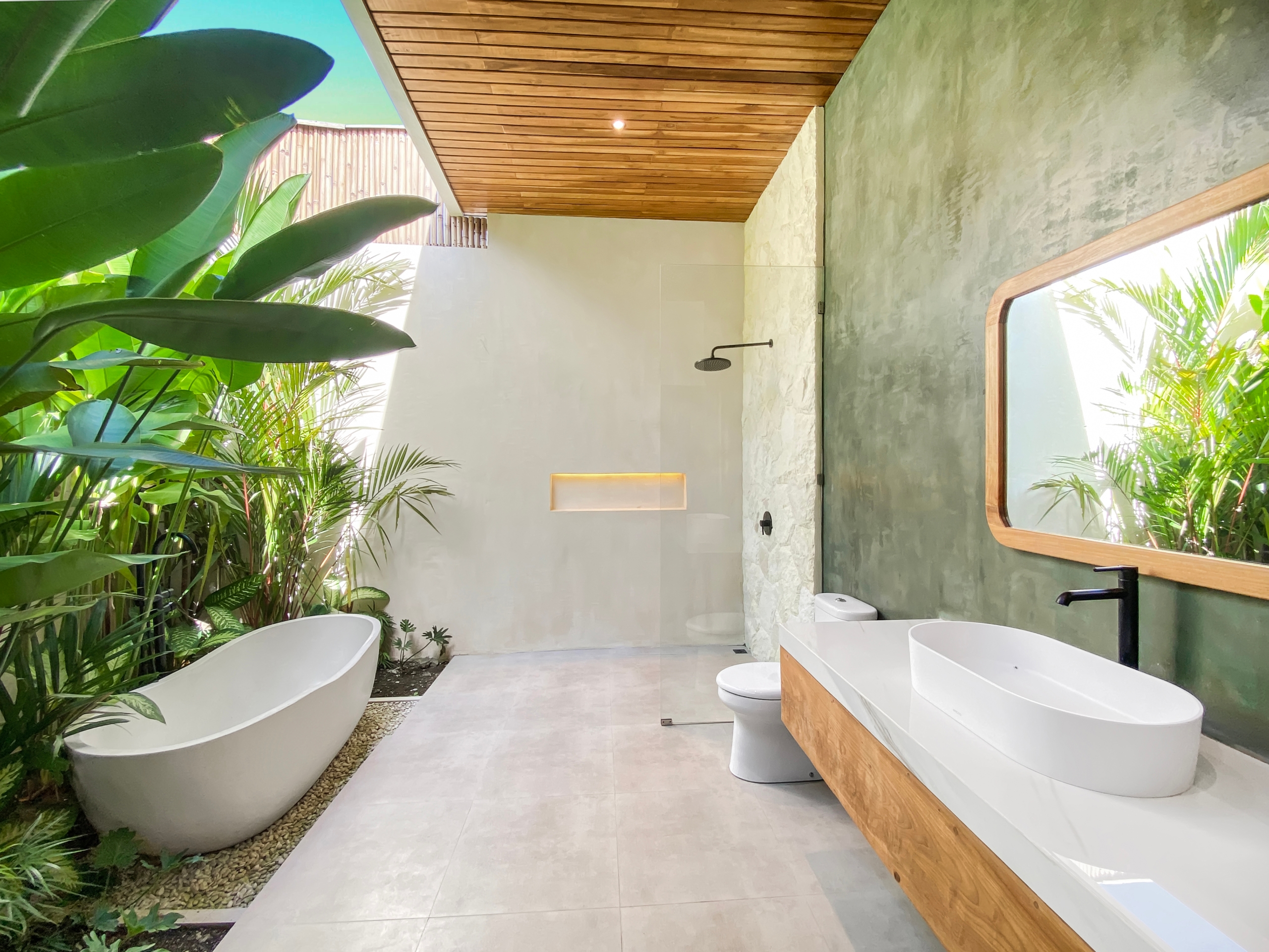 Modern bathroom with a white oval freestanding tub on a wooden base, surrounded by lush tropical plants and a light gray tiled floor.