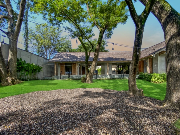 Courtyard with a pebble path, green lawn, and large trees; string lights hang overhead and a house with open doors is in the background.