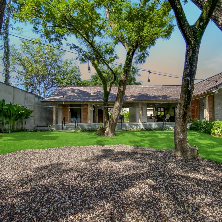 Courtyard with a pebble path, green lawn, and large trees; string lights hang overhead and a house with open doors is in the background.