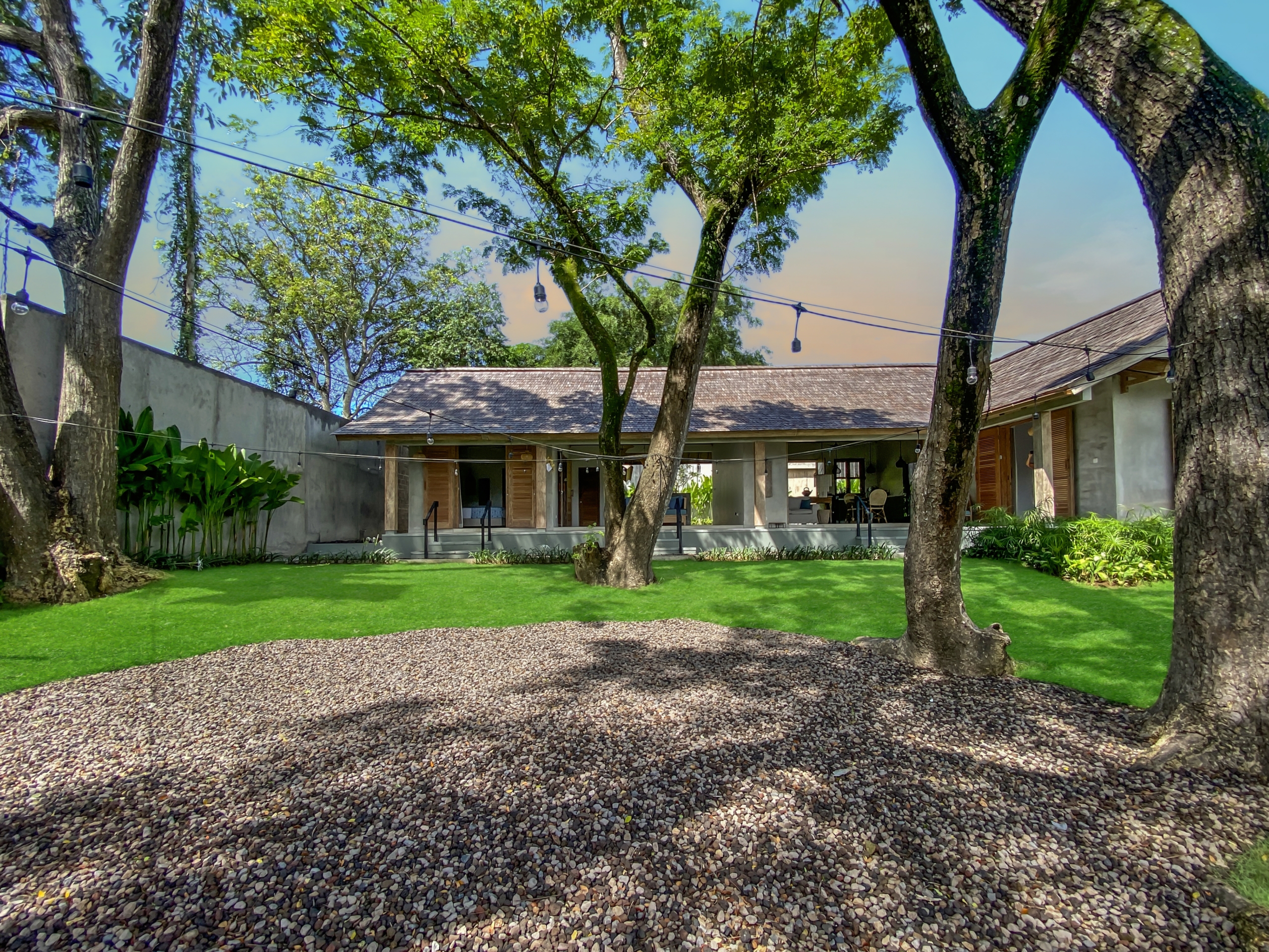 2b054e3ace69d1005fa2e6c688bb77f4817a583c Courtyard with a pebble path, green lawn, and large trees; string lights hang overhead and a house with open doors is in the background.