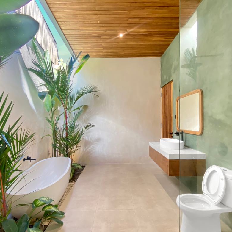 Modern bathroom with a white freestanding tub, tall tropical plants, and a wooden slat ceiling; light green walls and natural light.