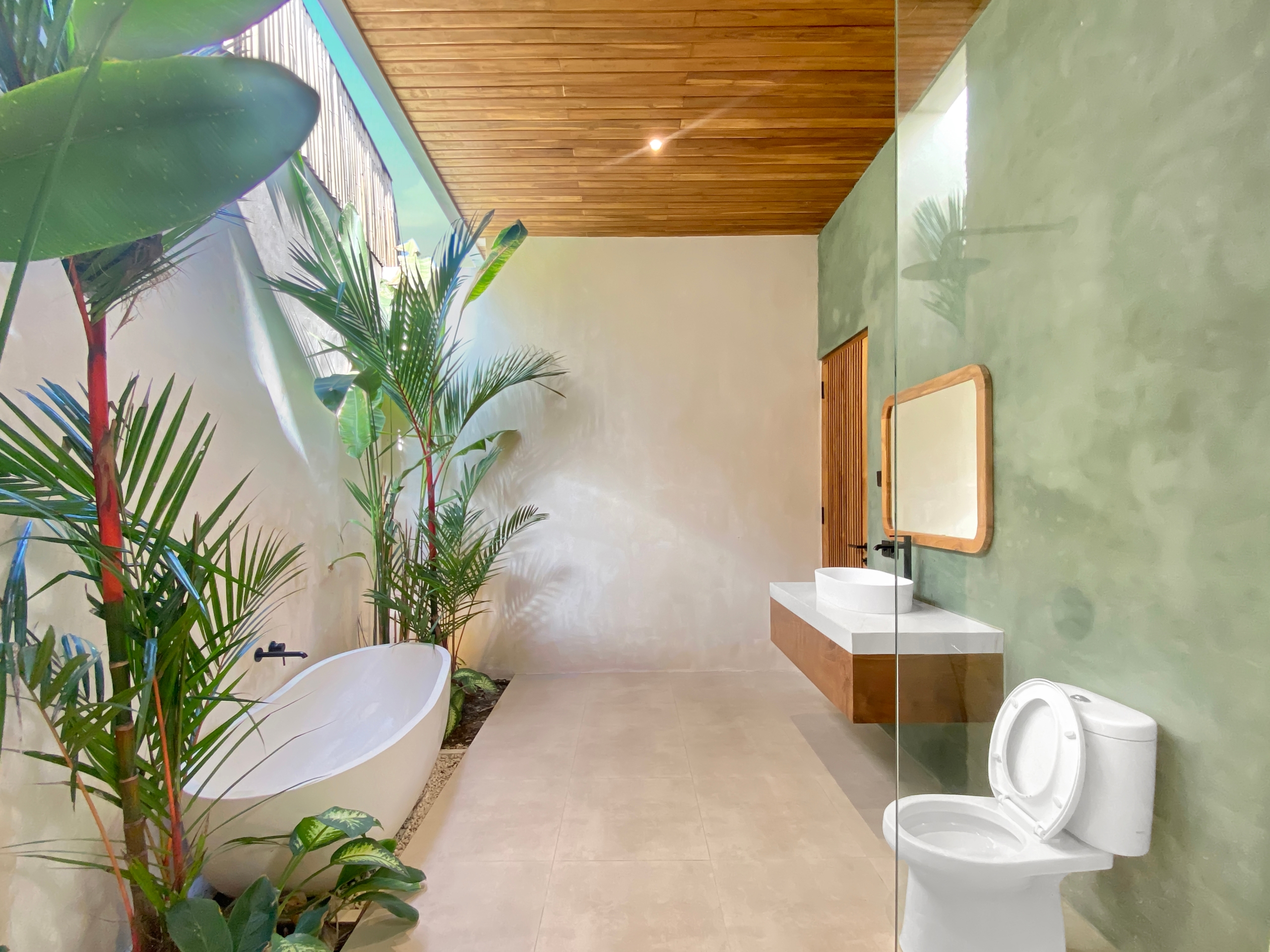 Modern bathroom with a white freestanding tub, tall tropical plants, and a wooden slat ceiling; light green walls and natural light.